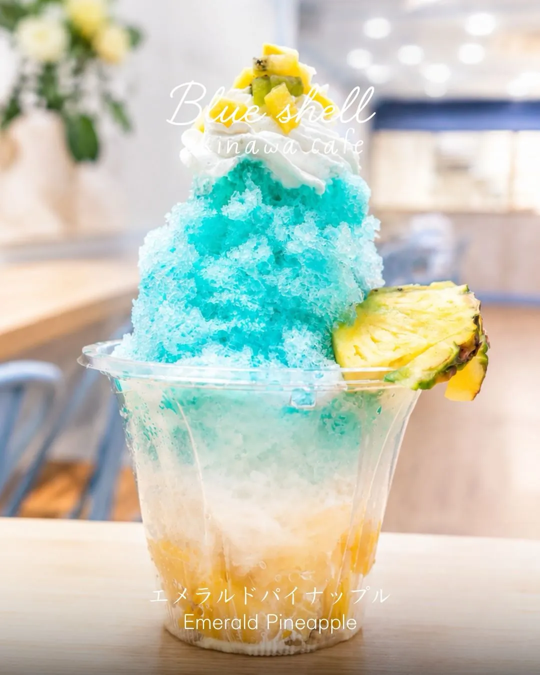 透明カップに入った水色と黄色のグラデーションのかき氷。ホイップクリームやパイナップルスライスがトッピングされている。「Blue shell Okinawa cafe」「エメラルドパイナップル Emerald Pineapple」の文字。