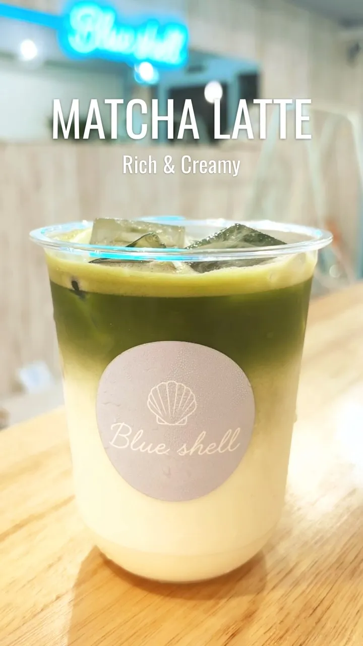 「MATCHA LATTE Rich & Creamy」の文字とともに、木目調のテーブルに置かれたアイス抹茶ラテの画像。透明カップには貝殻マークと「Blue shell」と書かれた丸いシールが貼られ、白と緑の2層になっています。