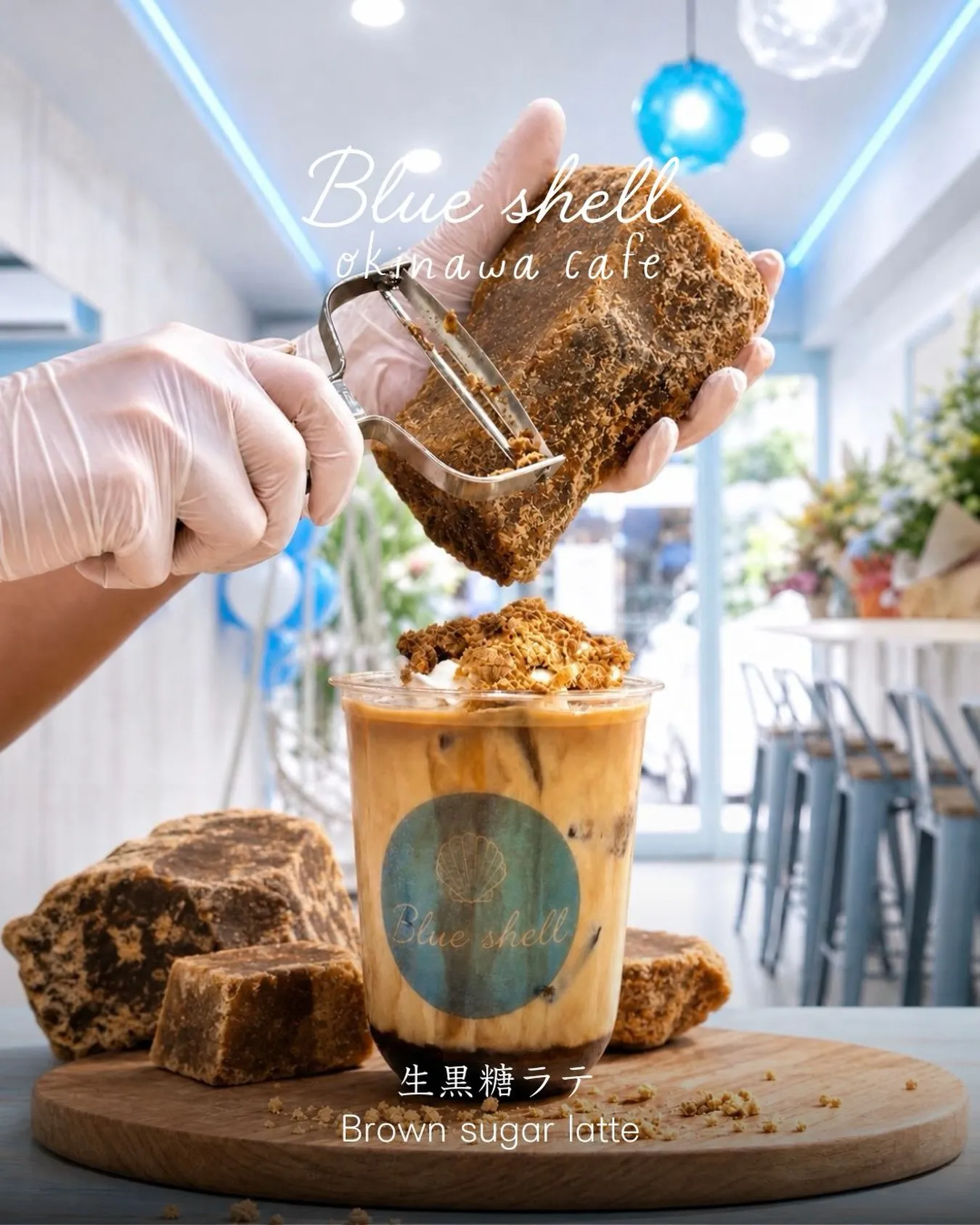 カフェ店内で、白い手袋をはめた手が黒糖の塊をピーラーで削り、手前の生黒糖ラテにトッピングしている様子。上部に「Blue shell okinawa cafe」、下部に「生黒糖ラテ Brown sugar latte」の文字。