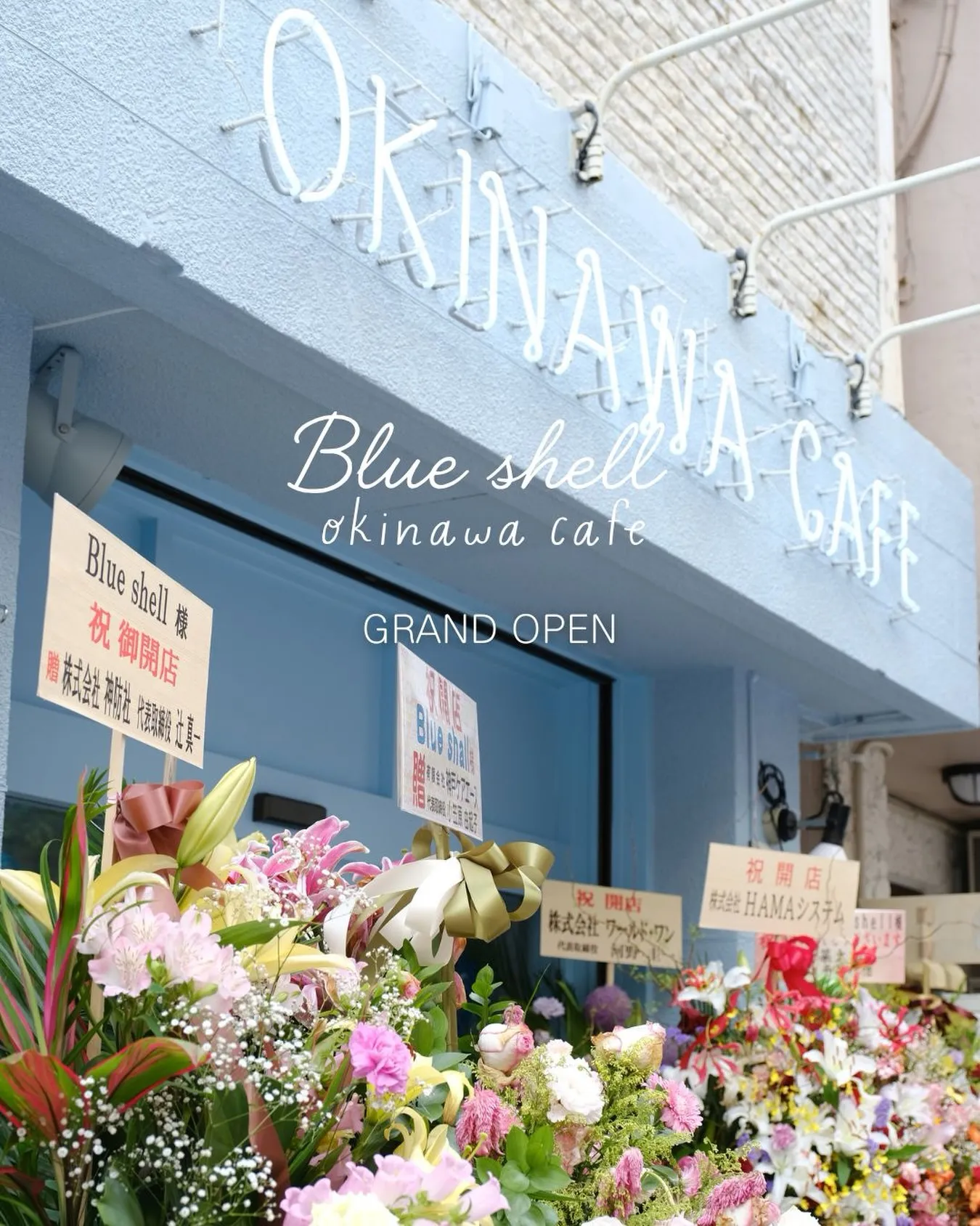 淡い水色の外壁に「OKINAWA CAFE」の看板が掲げられた店舗の前に、色とりどりの開店祝いの花スタンドが並ぶ様子。中央に「Blue shell okinawa cafe GRAND OPEN」、祝い札に「祝 御開店」「Blue shell 様」などの文字が写っている。