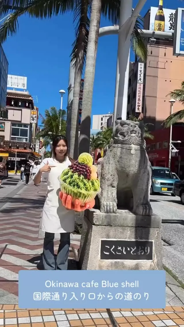 国際通りのシーサー像（台座に「こくさいどおり」）の横で、白い服を着た女性が巨大な抹茶かき氷を指差す様子。下部には「Okinawa cafe Blue shell 国際通り入り口からの道のり」の文字。
