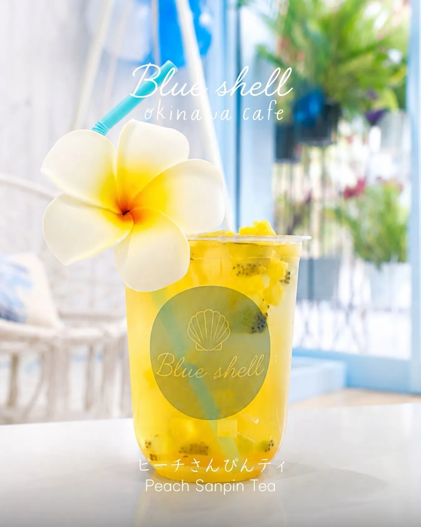 プルメリアの花と水色のストローが添えられた、カットフルーツ入りのピーチさんぴん茶。カップには「Blue shell」のロゴがあり、画像上部に「Blue shell okinawa cafe」、下部に「ピーチさんぴんティ Peach Sanpin Tea」と記載されている。