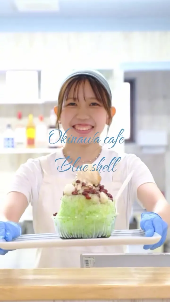 白い服とエプロン姿の笑顔の女性が、抹茶と小豆のかき氷が載ったトレイを差し出している様子。中央に「Okinawa cafe Blue shell」の文字。