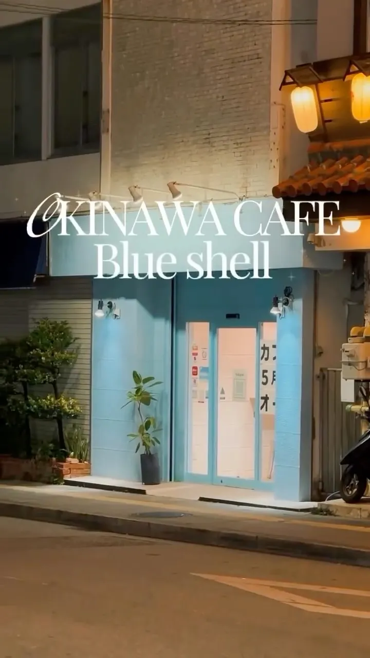 夜の水色のカフェ店舗の外観。中央に「OKINAWA CAFE Blue shell」と白文字が表示され、ガラス戸には「カ」「5月」「オ」などの文字が見える。