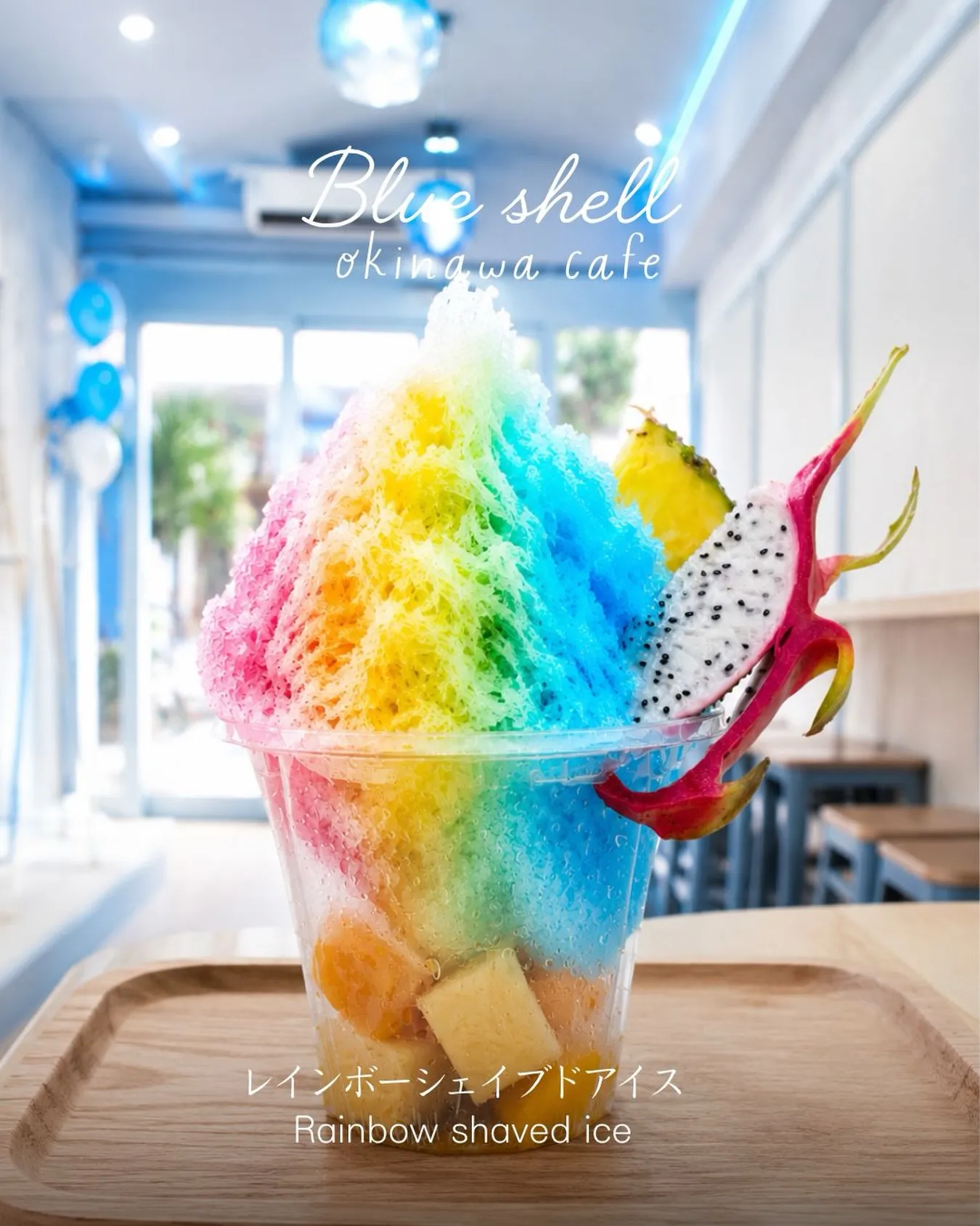 「Blue shell okinawa cafe」の「レインボーシェイブアイス Rainbow shaved ice」。青と白のカフェ店内にて、木製トレーに置かれたカラフルなシロップのかき氷。ドラゴンフルーツとパイナップルが添えられている。