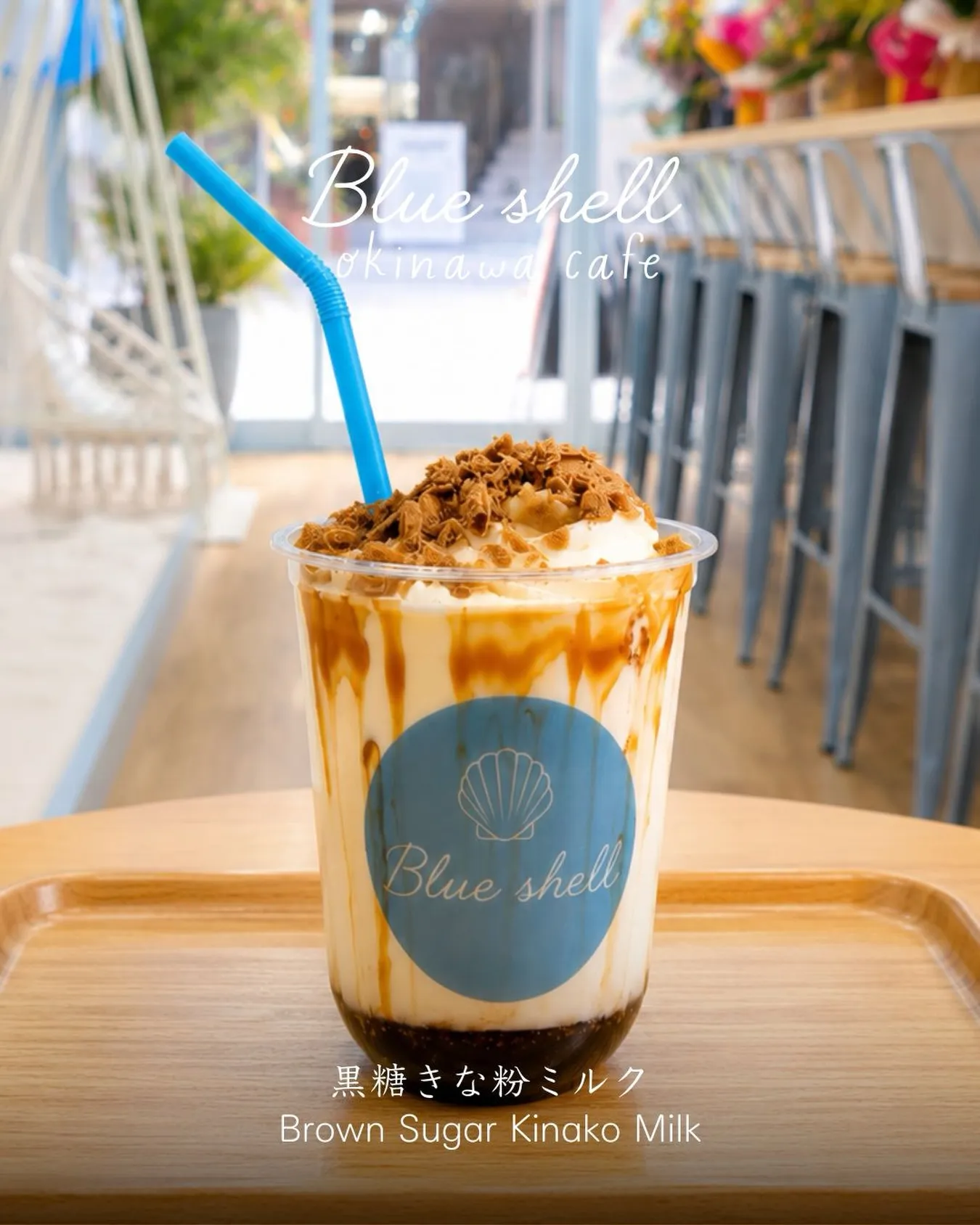 カフェのトレイに置かれた「黒糖きな粉ミルク(Brown Sugar Kinako Milk)」。貝殻ロゴと「Blue shell」のシール付き透明カップに、黒糖シロップ、ミルク、ホイップ、フレーク状トッピング、青いストローが入っている。上部に「Blue shell okinawa cafe」の文字。