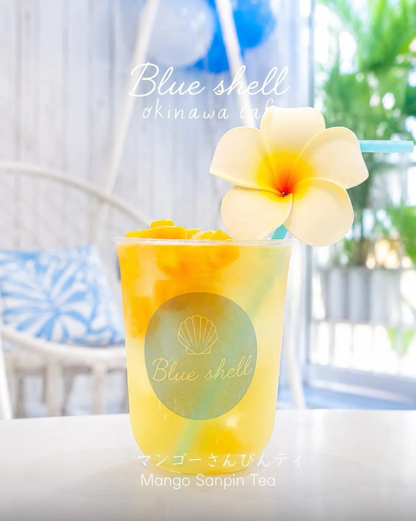 プルメリアの花とストローが添えられたマンゴーさんぴんティのドリンクカップ。「Blue shell okinawa cafe」「Blue shell」「マンゴーさんぴんティ Mango Sanpin Tea」の文字入り。
