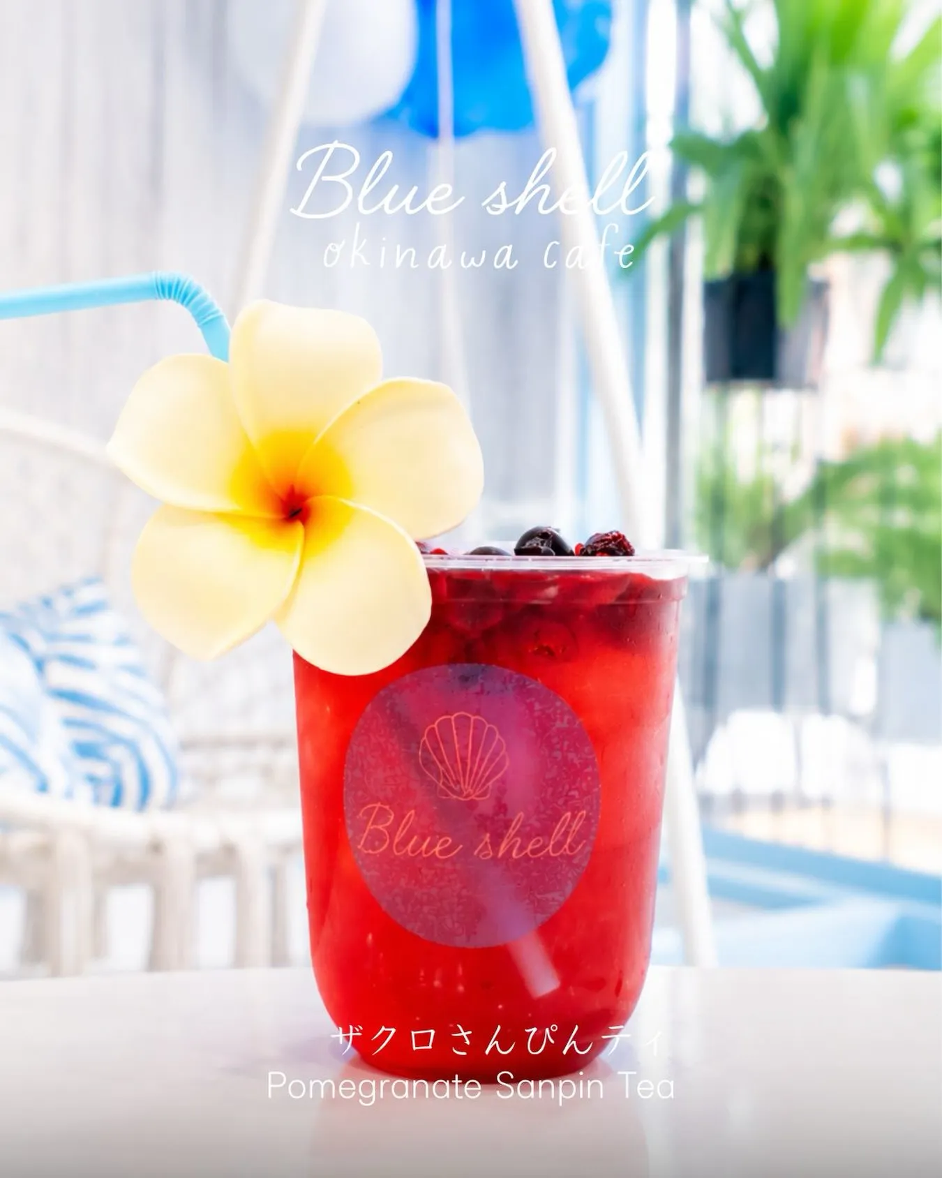 カフェのテラス席に置かれた、プルメリアの花と水色のストローが添えられた赤いドリンク「ザクロさんぴんティ(Pomegranate Sanpin Tea)」。カップには「Blue shell」のロゴがあり、画像上部にも「Blue shell okinawa cafe」の文字が記されています。