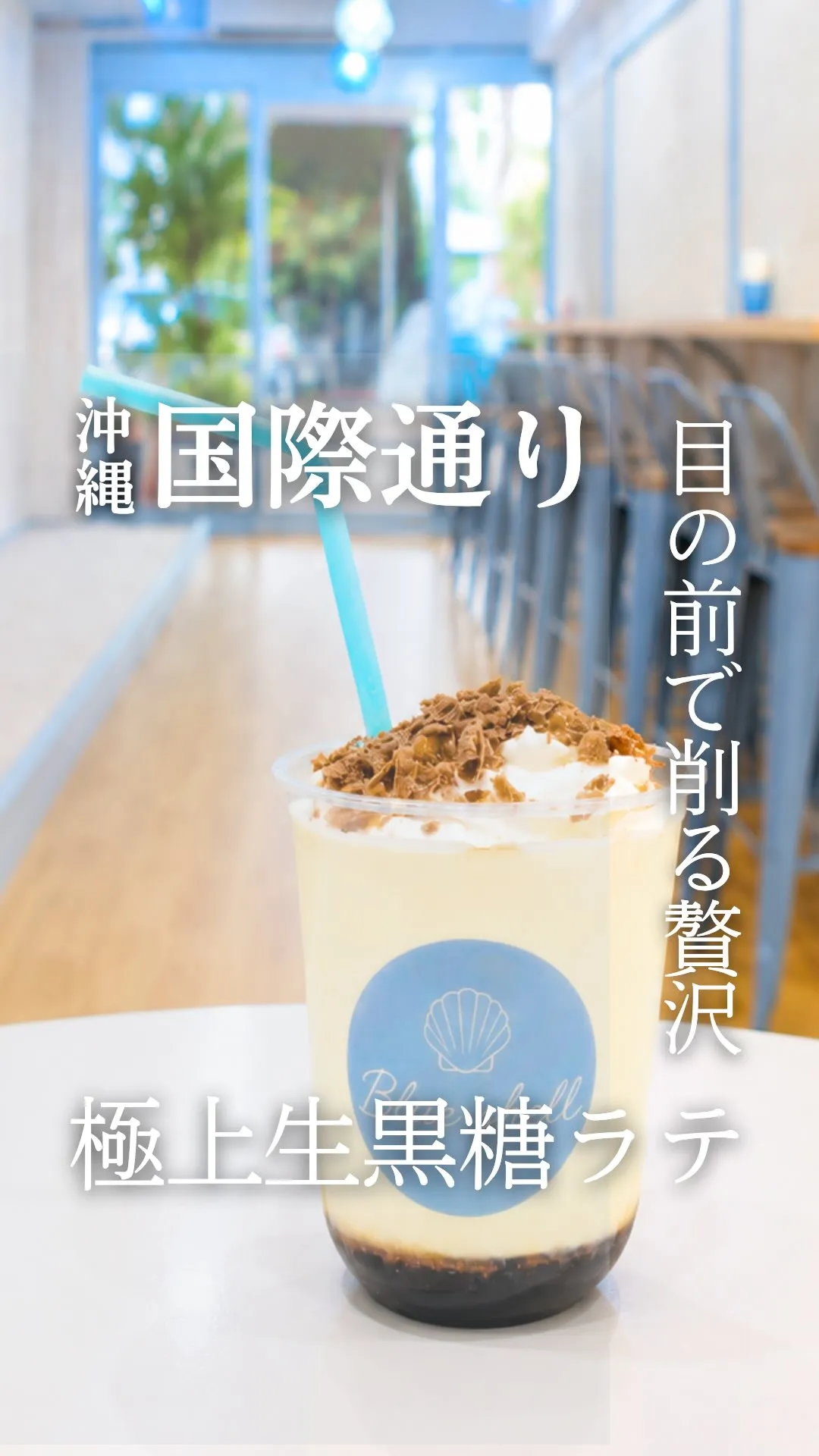 カフェのテーブルに置かれた「極上生黒糖ラテ」のカップ。底に黒糖、上にホイップクリームと削り黒糖が乗り、水色のストローが刺さっています。画像には「沖縄」「国際通り」「目の前で削る贅沢」「極上生黒糖ラテ」の文字が入っています。