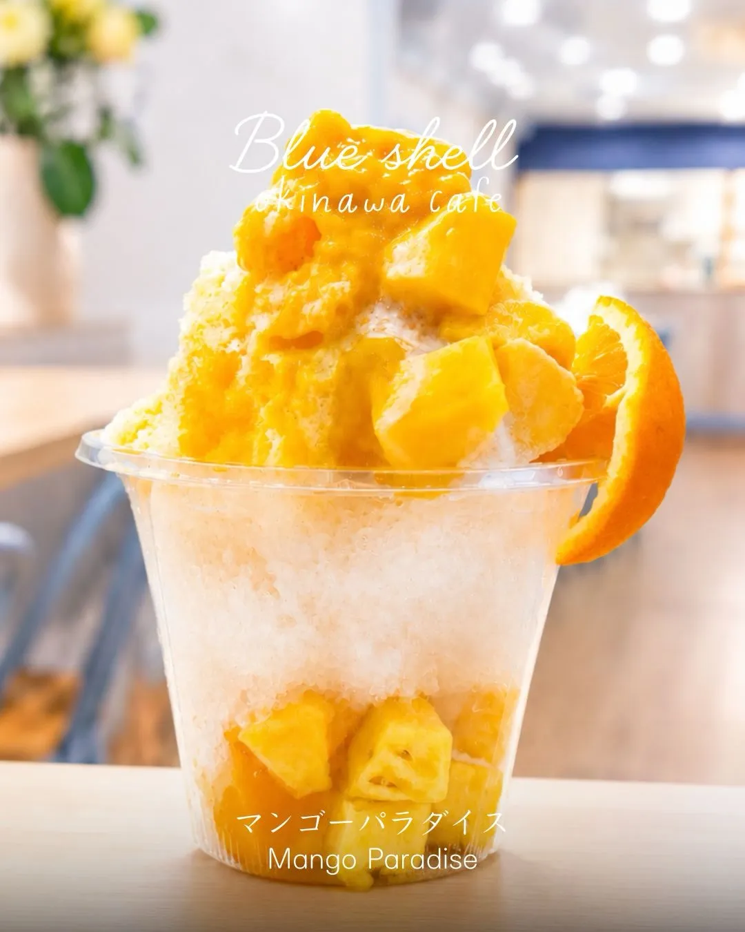 カフェ店内で撮影された、透明カップ入りのかき氷「マンゴーパラダイス Mango Paradise」。上部にマンゴー果肉とオレンジが乗り、底にも果肉が入っている。上部に「Blue shell Okinawa cafe」の文字。